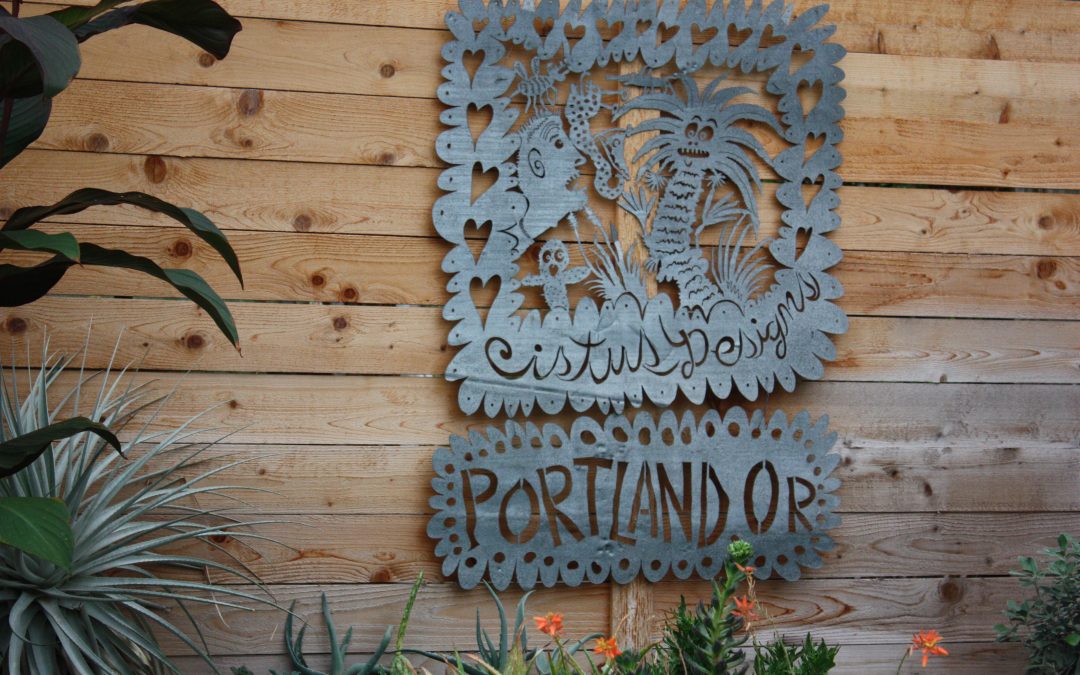 A metal sign hanging on a wooden nursery wall featuring fantastical palm and cactus plant and monster faces with the name Cistus Nursery, Portland, OR included at the bottom. You can see some plants on a table below the sign.